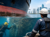 Underwater diver performing hull cleaning on a large commercial vessel in Singapore port while a marine engineer supervises maintenance operations above water.